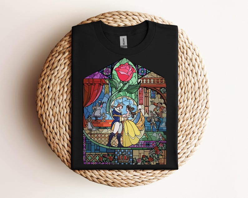 May include: Black t-shirt featuring a stained-glass design with characters from a fairy tale. The design includes a red rose, a couple dancing, and other characters. The shirt is folded on a woven surface.