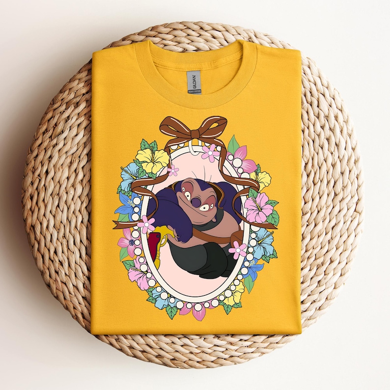 Puede incluir: Camiseta de color amarillo mostaza con un personaje de dibujos animados dentro de un marco ovalado. El marco est&aacute; decorado con un lazo marr&oacute;n, cintas y flores coloridas. El personaje tiene pelo morado y sonr&iacute;e.