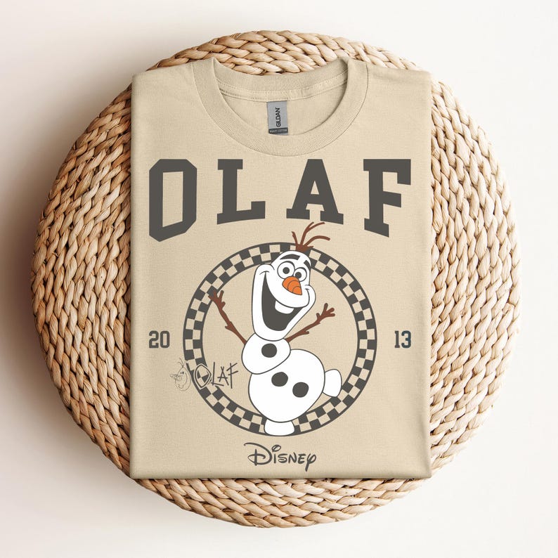 Może przedstawiać: Beżowa koszulka z czarno-białym kwadratowym kołem przedstawiającym Olafa z Krainy Lodu. Tekst "OLAF" znajduje się nad kołem, a "DISNEY" pod kołem. Liczby "20" i "13" znajdują się po obu stronach koła.