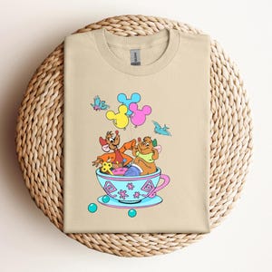 May include: Tan t-shirt featuring a colorful cartoon design. The graphic depicts two cartoon characters in a teacup with Mickey Mouse-shaped balloons. The design includes blue, pink, yellow, and green accents. The shirt is folded on a woven surface.