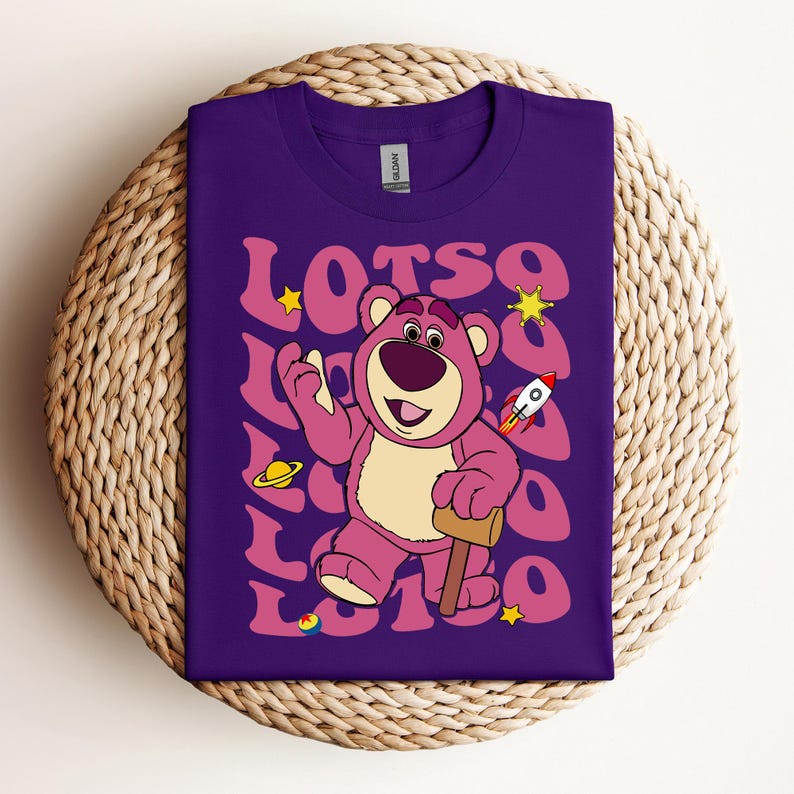 Puede incluir: Camiseta morada con un oso rosa de dibujos animados que lleva un cohete rojo y sostiene un martillo. El oso est&aacute; rodeado de la palabra "LOTSO" en una fuente retro.