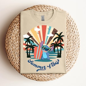 May include: A tan t-shirt with a cartoon character wearing sunglasses and holding a surfboard. The design includes palm trees, a colourful sunset, and the words "Summer Vibes" in blue script. The shirt is folded on a woven surface.