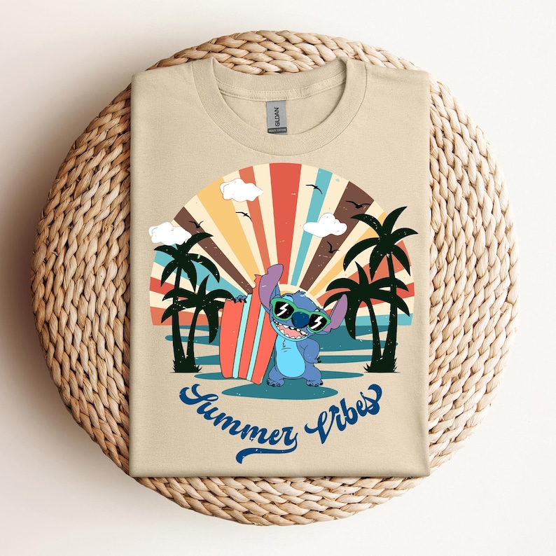May include: A tan t-shirt with a cartoon character wearing sunglasses and holding a surfboard. The design includes palm trees, a colourful sunset, and the words "Summer Vibes" in blue script. The shirt is folded on a woven surface.
