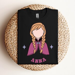 May include: A black t-shirt featuring a graphic illustration of a character with blonde hair in two braids, wearing a magenta cape with a blue clasp. The character has no facial features. Two light blue four-pointed stars and the text ANNA are printed.
