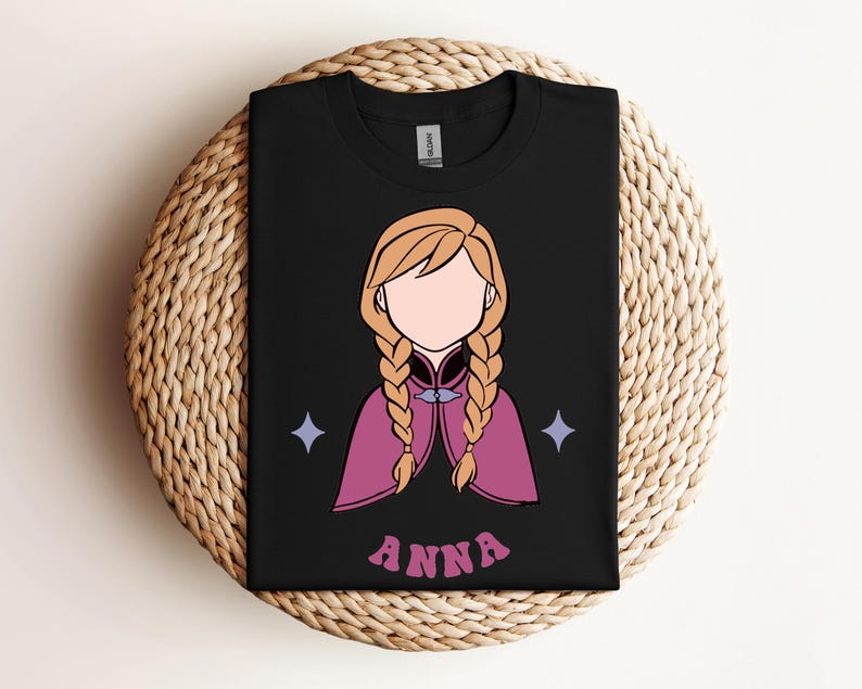 May include: A black t-shirt featuring a graphic illustration of a character with blonde hair in two braids, wearing a magenta cape with a blue clasp. The character has no facial features. Two light blue four-pointed stars and the text ANNA are printed.