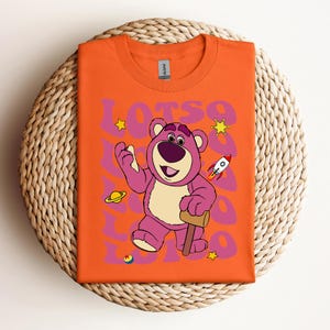 Puede incluir: Camiseta naranja con un oso de dibujos animados rosa que lleva una camisa azul y sostiene un martillo. El oso est&aacute; rodeado de estrellas y el texto "Lots-o".