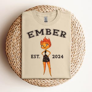 May include: A beige t-shirt with the text "EMBER EST. 2024" printed in black. A cartoon character with orange hair and a black dress is in the center of the shirt.