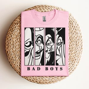 May include: A light pink t-shirt featuring a graphic of four cartoon villains in black and white vertical panels. The text "BAD BOYS" is printed in black block letters below the panels. The shirt is folded on a woven, circular surface.