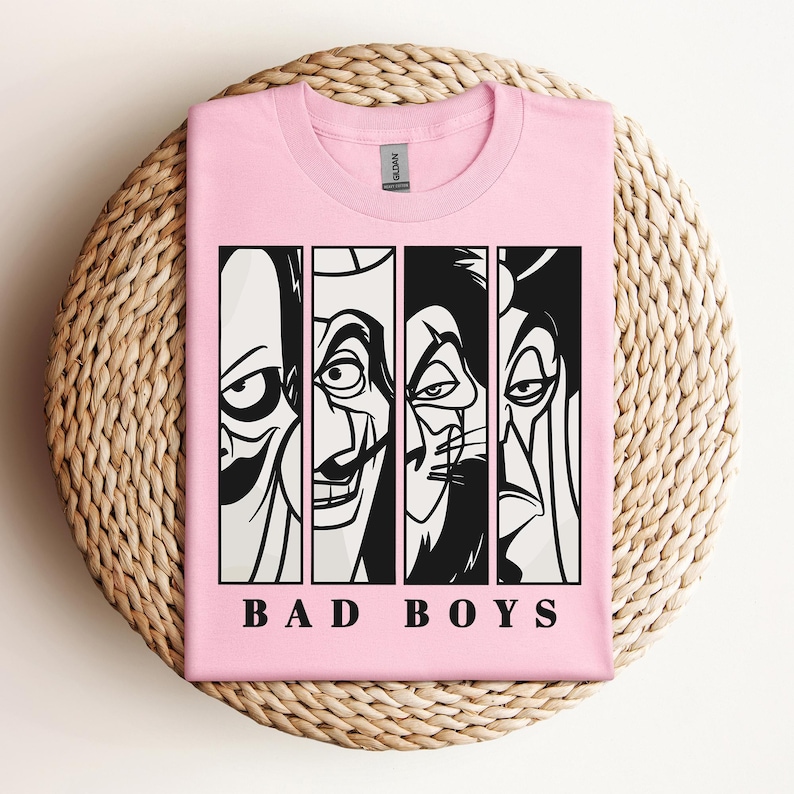 May include: A light pink t-shirt featuring a graphic of four cartoon villains in black and white vertical panels. The text "BAD BOYS" is printed in black block letters below the panels. The shirt is folded on a woven, circular surface.