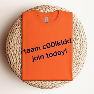 May include: An orange t-shirt with the text "team c00lkidd join today!" in black. The shirt is folded and placed on a round, woven, beige surface. The Gildan brand label is visible on the collar.