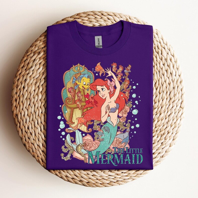 May include: A purple t-shirt featuring a graphic of Ariel from The Little Mermaid. The design includes Ariel, Flounder, and other characters, with the words "The Little Mermaid" in teal. The shirt is displayed on a woven surface.