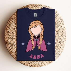 May include: A navy blue t-shirt featuring a graphic illustration of a character with blonde hair in two braids, wearing a magenta cape. The character has no facial features. Two light purple four-pointed stars flank the figure, and the name ANNA is printed in pink letters below.