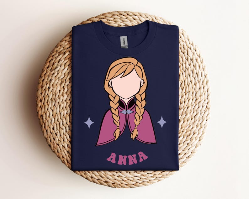 May include: A navy blue t-shirt featuring a graphic illustration of a character with blonde hair in two braids, wearing a magenta cape. The character has no facial features. Two light purple four-pointed stars flank the figure, and the name ANNA is printed in pink letters below.