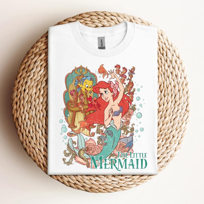 May include: White t-shirt featuring a colorful graphic of Ariel from The Little Mermaid. The design includes Ariel with red hair, a blue seashell bra, and a turquoise tail, surrounded by sea creatures and the words "The Little Mermaid."