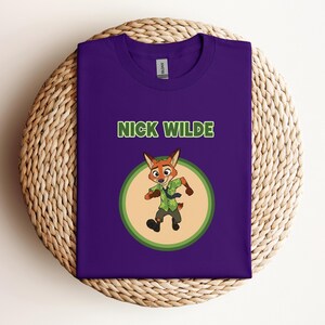 May include: A purple t-shirt featuring the text "NICK WILDE" in green letters above a cartoon image of a fox character. The fox is wearing a green shirt and tie, set within a circular design with a light green and tan border.