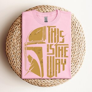 May include: A pink t-shirt with a gold graphic of a helmet and the text "THIS IS THE WAY." The shirt is folded and placed on a woven, circular placemat. The design has a distressed, vintage look.