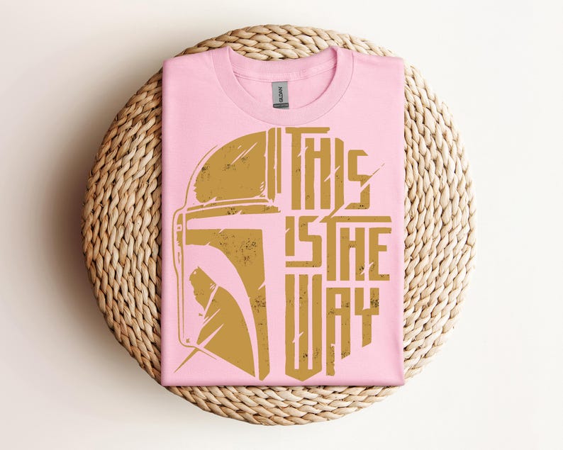 May include: A pink t-shirt with a gold graphic of a helmet and the text "THIS IS THE WAY." The shirt is folded and placed on a woven, circular placemat. The design has a distressed, vintage look.
