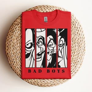 May include: A red t-shirt featuring a graphic of four cartoon villains in black and white, with the words "BAD BOYS" in a bold font at the bottom. The shirt is folded on a woven, circular surface.