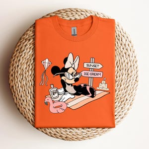 K&ouml;nnte beinhalten: Orangefarbenes T-Shirt mit einem Cartoon-Bild von Minnie Mouse, die eine Sonnenbrille und eine Schleife tr&auml;gt und sich auf einem Strandtuch mit einem rosafarbenen Flamingo-Schwimmtier, einer Flasche Sonnencreme und einem Schild mit der Aufschrift "Sunset" und "Ice-Cream" entspannt.