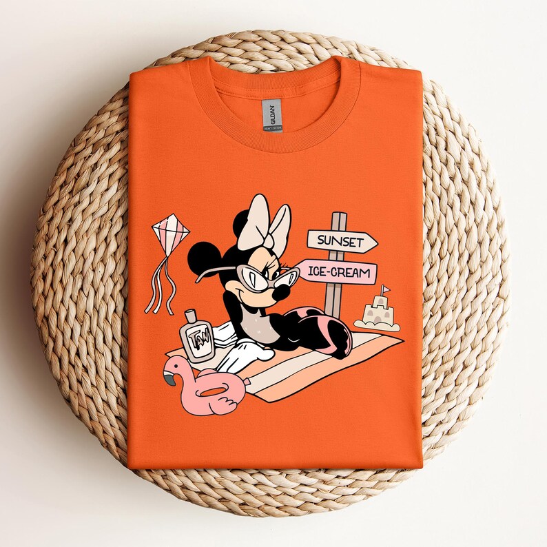 K&ouml;nnte beinhalten: Orangefarbenes T-Shirt mit einem Cartoon-Bild von Minnie Mouse, die eine Sonnenbrille und eine Schleife tr&auml;gt und sich auf einem Strandtuch mit einem rosafarbenen Flamingo-Schwimmtier, einer Flasche Sonnencreme und einem Schild mit der Aufschrift "Sunset" und "Ice-Cream" entspannt.