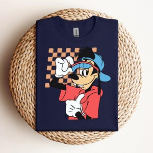 May include: A navy blue t-shirt featuring a cartoon image of Goofy wearing a red shirt, blue hat, and sunglasses. The background is a checkered pattern in orange and brown.
