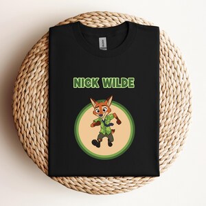 May include: Black t-shirt featuring a cartoon image of Nick Wilde from Zootopia. The design includes the character in a green and brown outfit inside a green and tan circle, with the name "NICK WILDE" above. The shirt is displayed on a woven surface.