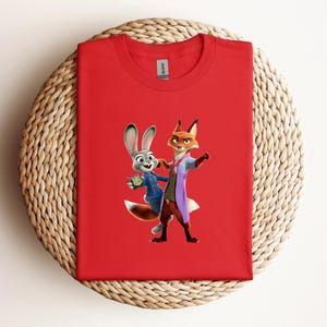 May include: A red t-shirt featuring a cartoon image of Judy Hopps and Nick Wilde from Zootopia. Judy, a gray rabbit, is wearing a blue uniform. Nick, a fox, is wearing a purple coat. The shirt is made of a soft material.