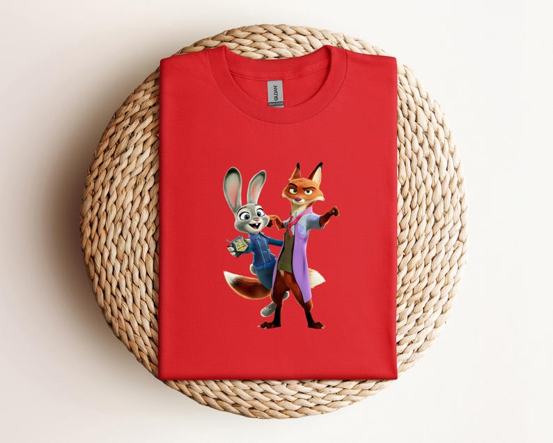 May include: A red t-shirt featuring a cartoon image of Judy Hopps and Nick Wilde from Zootopia. Judy, a gray rabbit, is wearing a blue uniform. Nick, a fox, is wearing a purple coat. The shirt is made of a soft material.