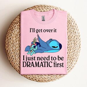 Puede incluir: Camiseta rosa con un personaje azul Stitch de Lilo & Stitch. Stitch est&aacute; acostado con una peque&ntilde;a mu&ntilde;eca. El texto en la camiseta dice "I'll get over it I just need to be DRAMATIC first."