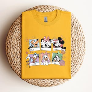 May include: A mustard yellow crew neck t-shirt featuring a graphic print of six Polaroid-style frames. Each frame contains a Disney character illustration with their name written below: Pluto, Minnie, Mickey, Donald, Daisy, and Goofy.