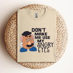 May include: A beige t-shirt with the text "DON'T MAKE ME USE MY ANGRY EYES". The design includes a cartoon character with a black hat, a brown mustache, and blue shoes. The t-shirt is folded on a circular woven mat, likely made of wicker or straw.