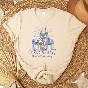 Puede incluir: Camiseta beige con una ilustraci&oacute;n colorida de un castillo de cuento de hadas con el texto "Florida!!!" y "one hell of a drug." El castillo es azul, morado y dorado, con fuegos artificiales y corazones. La camiseta es de material suave.