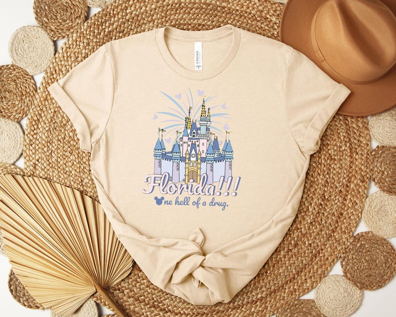 Puede incluir: Camiseta beige con una ilustraci&oacute;n colorida de un castillo de cuento de hadas con el texto "Florida!!!" y "one hell of a drug." El castillo es azul, morado y dorado, con fuegos artificiales y corazones. La camiseta es de material suave.