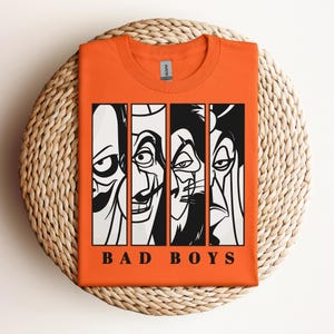 May include: Orange t-shirt featuring a graphic of four cartoon villains in black and white vertical panels. The text "BAD BOYS" is printed in black below the panels. The shirt is displayed on a woven, circular surface.