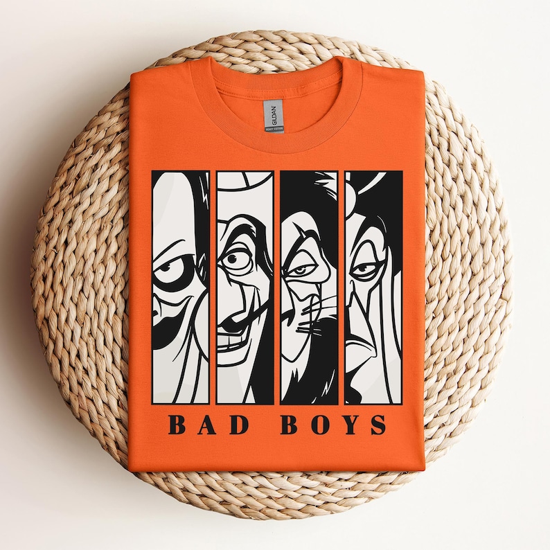 May include: Orange t-shirt featuring a graphic of four cartoon villains in black and white vertical panels. The text "BAD BOYS" is printed in black below the panels. The shirt is displayed on a woven, circular surface.