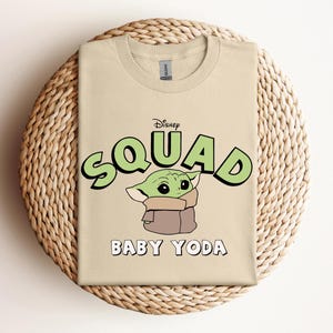 May include: A tan t-shirt featuring the text "Disney Squad" in green with a black outline, and the words "Baby Yoda" in white. The shirt has a graphic of Baby Yoda. The t-shirt is displayed on a woven, circular placemat.