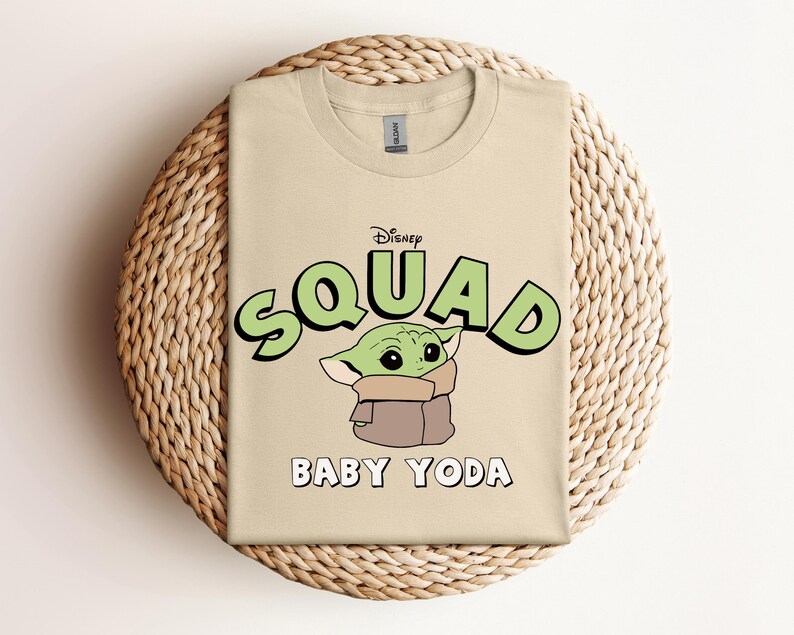 May include: A tan t-shirt featuring the text "Disney Squad" in green with a black outline, and the words "Baby Yoda" in white. The shirt has a graphic of Baby Yoda. The t-shirt is displayed on a woven, circular placemat.