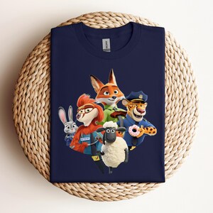 May include: Navy blue t-shirt featuring a colorful graphic of characters from the animated movie Zootopia. The design includes a fox, a rabbit, a sheep, a cheetah, and other characters. The shirt is displayed on a woven, circular surface.
