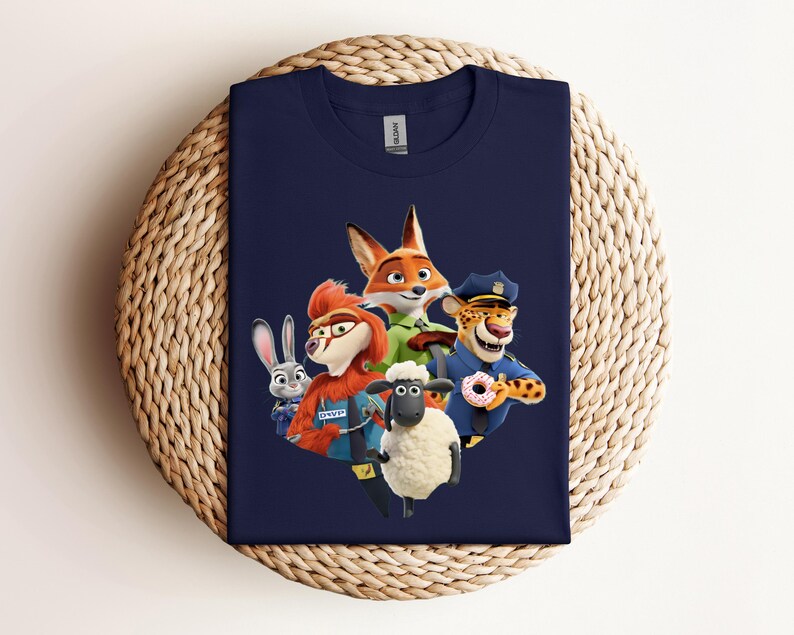 May include: Navy blue t-shirt featuring a colorful graphic of characters from the animated movie Zootopia. The design includes a fox, a rabbit, a sheep, a cheetah, and other characters. The shirt is displayed on a woven, circular surface.