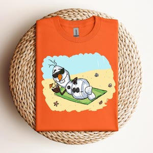 Op de afbeelding: Oranje T-shirt met een cartoon afbeelding van Olaf uit Frozen, die een zonnebril draagt en ontspant op een strandhanddoek met een tropisch drankje.