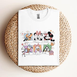 May include: A white crew neck t-shirt featuring a graphic print of six colorful polaroid-style frames. Each frame contains a cartoon character: Pluto, Minnie, Mickey, Donald, Daisy, and Goofy, with their names written in cursive script below.