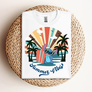 May include: White t-shirt with a cartoon character wearing sunglasses and holding a surfboard, set against a retro beach scene with palm trees and a colourful sunset. The text "Summer Vibes" is written in blue script.