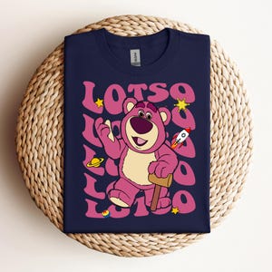 Puede incluir: Una camiseta azul marino con un oso de dibujos animados rosa que lleva un cohete rojo y sostiene un mazo. El oso est&aacute; rodeado de la palabra "LOTSO" en una fuente retro con estrellas y planetas.