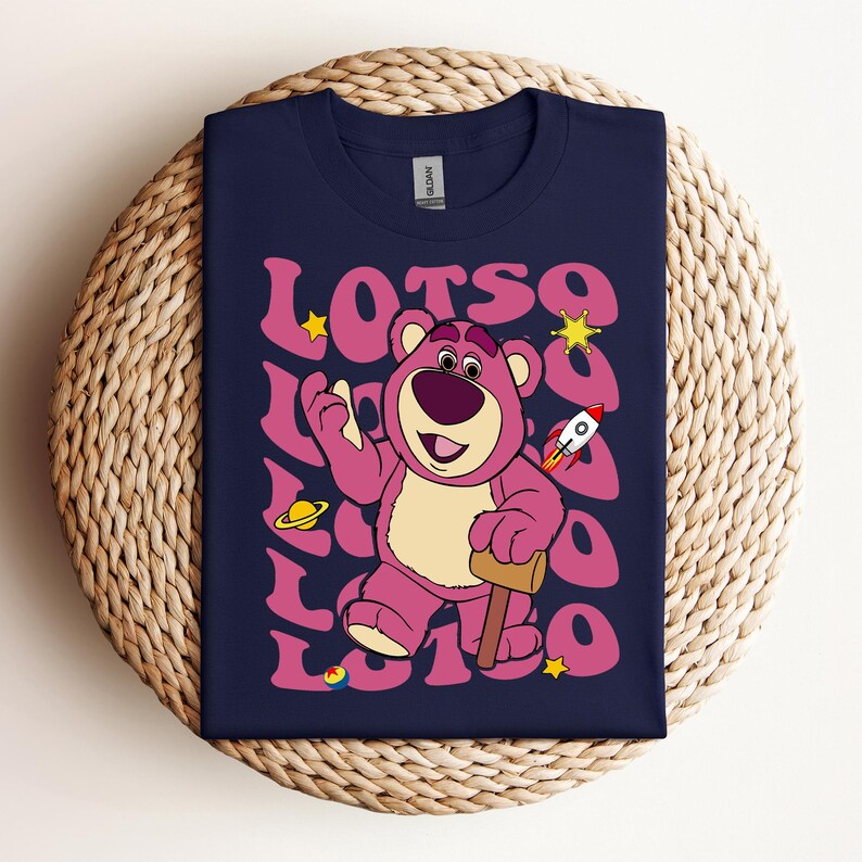 Puede incluir: Una camiseta azul marino con un oso de dibujos animados rosa que lleva un cohete rojo y sostiene un mazo. El oso est&aacute; rodeado de la palabra "LOTSO" en una fuente retro con estrellas y planetas.
