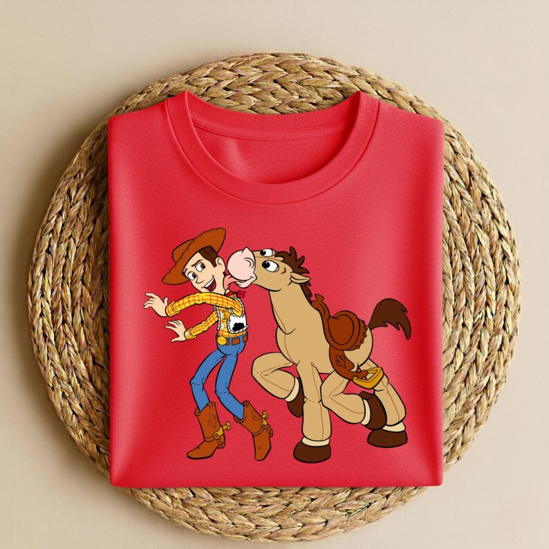Peut inclure: Un t-shirt rouge avec une illustration de dessin anim&eacute; d'un cow-boy et d'un cheval. Le cow-boy porte un chapeau marron, une chemise jaune, un jean bleu et des bottes marron. Le cheval est marron avec une selle.