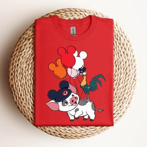 Op de afbeelding: Rood T-shirt met een cartoonvarken met een hoed, een haan en ballonnen. Het varken is wit met zwarte vlekken en een blauwe hoed. De haan is groen, geel en blauw. De ballonnen zijn rood, wit en oranje.