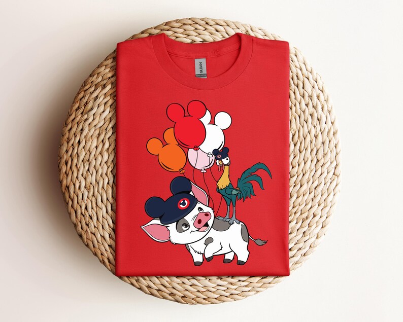 Op de afbeelding: Rood T-shirt met een cartoonvarken met een hoed, een haan en ballonnen. Het varken is wit met zwarte vlekken en een blauwe hoed. De haan is groen, geel en blauw. De ballonnen zijn rood, wit en oranje.
