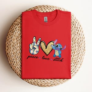 以下が含まれることがあります： リロ＆スティッチのスティッチが描かれた赤いTシャツ。ピースサイン、ハート、そして「peace love stitch」の文字が書かれています。