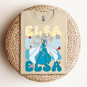Op de afbeelding: Beige T-shirt met een afbeelding van Elsa uit Frozen. Het ontwerp bevat de naam "ELSA" in een retro lettertype, met sneeuwvlokken en herfstbladeren. Elsa is afgebeeld in een blauwe jurk en witte cape.