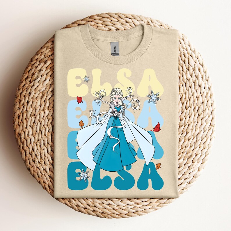 Op de afbeelding: Beige T-shirt met een afbeelding van Elsa uit Frozen. Het ontwerp bevat de naam "ELSA" in een retro lettertype, met sneeuwvlokken en herfstbladeren. Elsa is afgebeeld in een blauwe jurk en witte cape.